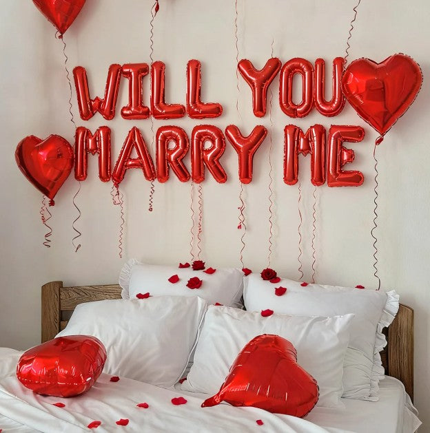Red 'WILL YOU MARRY ME' balloons above a bed with heart-shaped balloons and rose petals.
